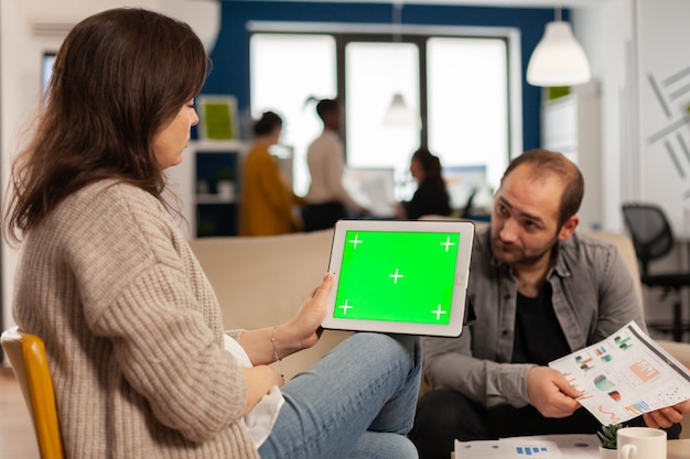 Back view of businesswoman sitting on couch using tablet with green screen talking with colleague while team working on background