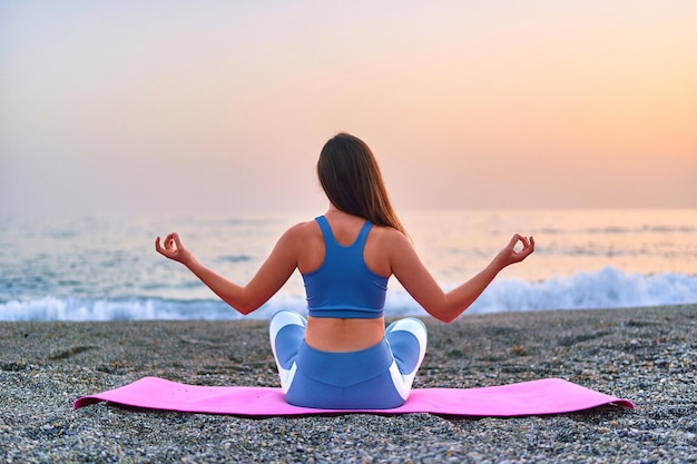 Back view of calm serene bliss satisfied fitness woman doing\
yoga meditation and breathing exercises on the beach by the sea\
mental mind care and healthy habits