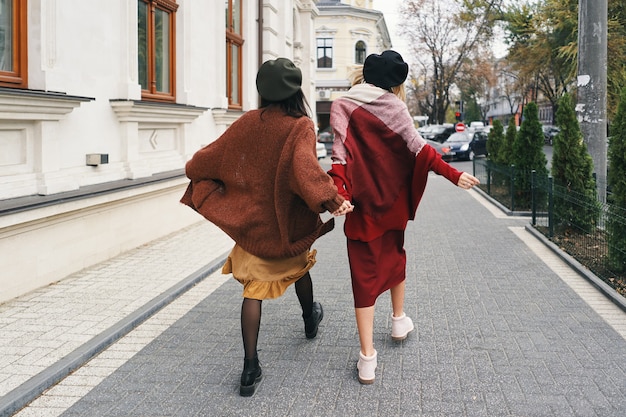 Back view. Carefree fashionable women models in elegant autumn clothes and glasses. Young fashion girls in stylish woven wool clothes and hats over urban city, autumn portrait.