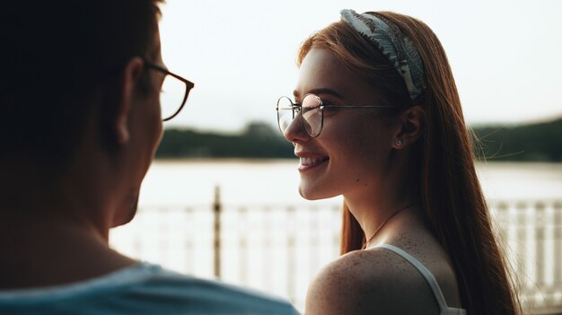 Back view of caucasian couple with eyeglasses sitting on a bridge looking at each other with a lake on background