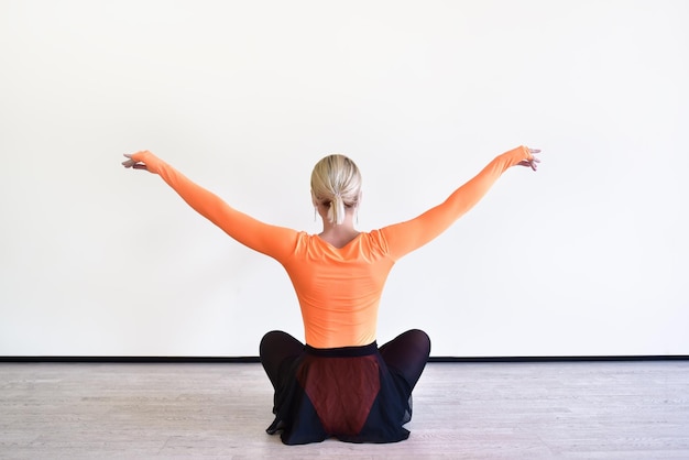 Back view of a charming blonde ballerina is sitting on the floor in the ballroom warming up before the performance