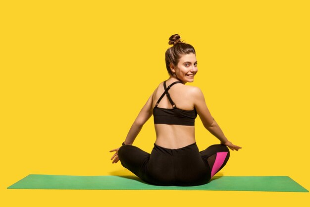 Back view cheerful woman with hair bun in tight sportswear sitting in lotus position on gym mat practicing yoga turning head to camera and smiling joyfully doing sports Happy healthy lifestyle