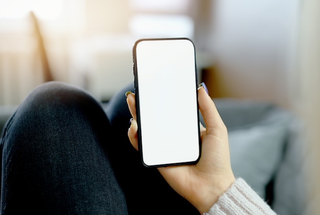 Back view close up of a woman hand holding and using smart phone with white blank empty screen sitting on a bed at home.