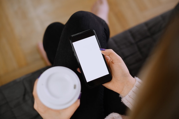 Vista posteriore ravvicinata di una mano di donna che tiene e utilizzando smart phone con schermo vuoto vuoto bianco seduto su un letto a casa.