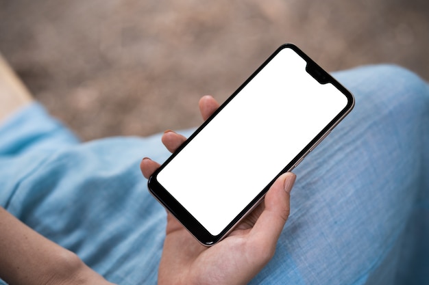 Photo back view close up of a woman hand using a smart phone with blank screen lying on a couch at home.