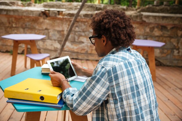 Back view of concenrated young man sitting and using tablet at the table