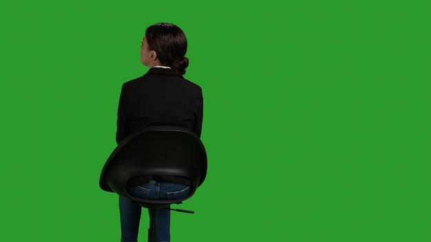 Back view of corporate worker sitting on chair and waiting on camera, wearing formal office suit and preparing in studio. Female model working as company employee over greenscreen backdrop.