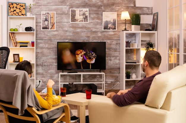 Photo back view of couple in living room watching a movie on the tv while eating takeaway food