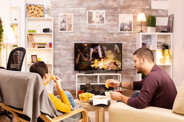 Back view of couple in living room watching a movie on the tv\
while eating takeaway food