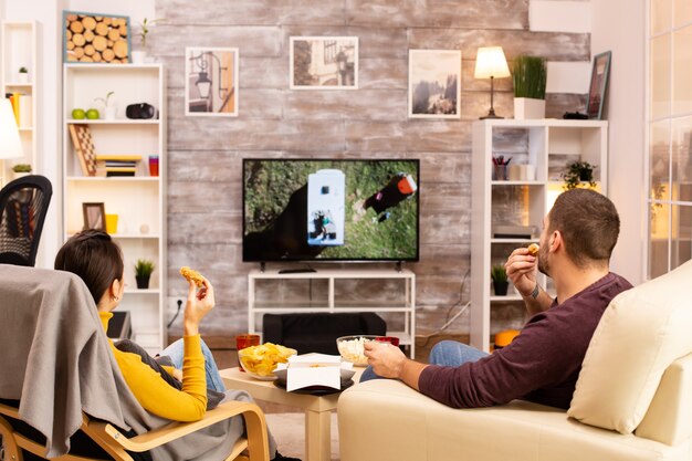 Photo back view of couple in living room watching a movie on the tv while eating takeaway food