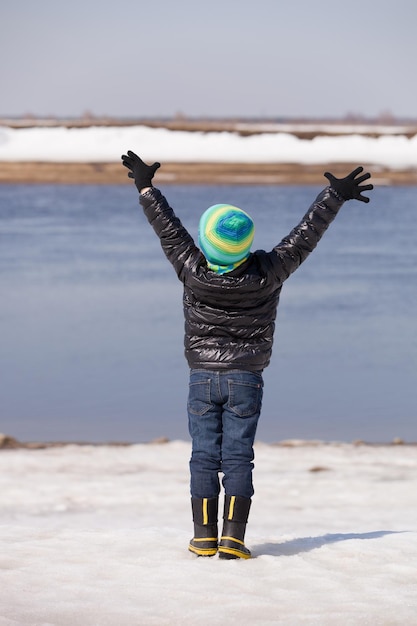 Back view on a cute happy kid boy standing on the river shore with his hands up on sunny winter day