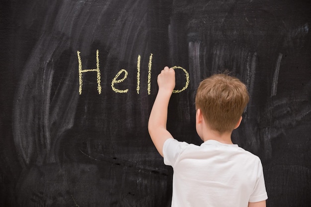 Vista posteriore sul ragazzo carino che scrive ciao parola con il gesso sul tabellone nella classe scolastica scuola elementare di istruzione