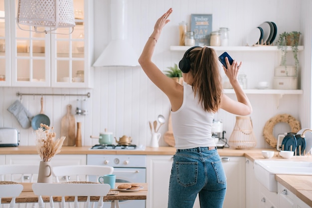 Back view on dancing beautiful Italian girl in headphones white vest and blue denim jeans listening woman rises hand holds phone Playful young hispanic woman enjoys weekend at home kitchen