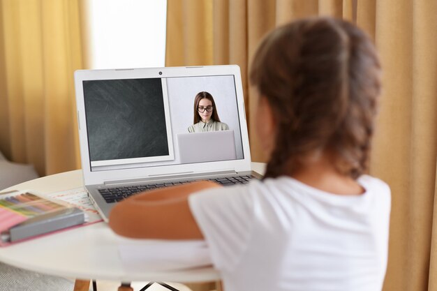 Back view of dark haired girl with pi tails sitting at desk at\
home on quarantine talk on video call with teacher, schoolgirl have\
online webcam conversation, engaged in web lesson.