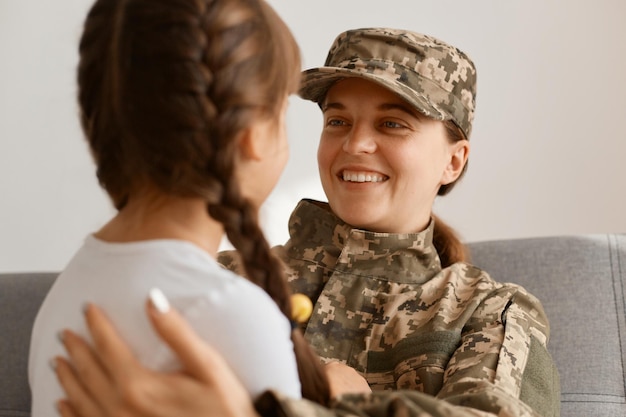 Back view of dark haired little kid with braids standing in\
front her mother woman wearing camouflage uniform and cap posing\
with her daughter military female arrived home from army