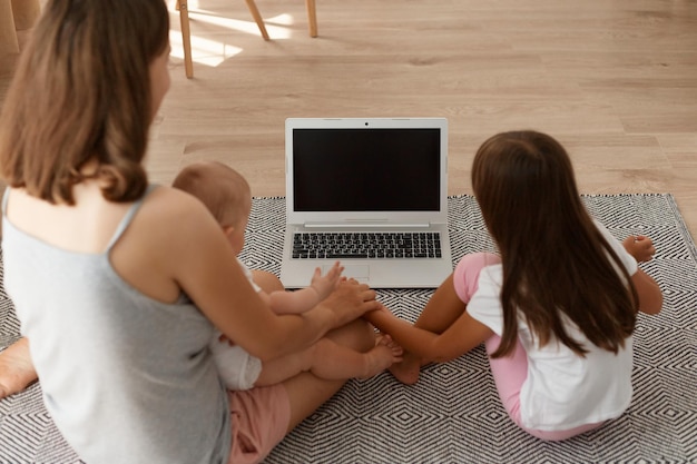 Back view of dark haired mother and daughter sitting on the living room floor and looking at laptop display, copy space for promotional text or your advertisement.