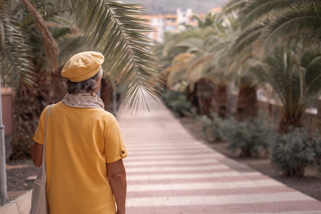Вид сзади на пожилую женщину в желтом с кепке, гуляющую в городском парке с пальмами