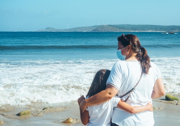 Back view of an embraced mother and daughter looking out to sea during the Coronavirus pandemic.