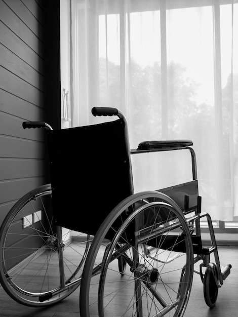 The back view of empty black wheelchair near wall with white\
curtain at glass window in the living room vertical style waiting\
for patient services lonely wheelchair with nobody in hospital\
room