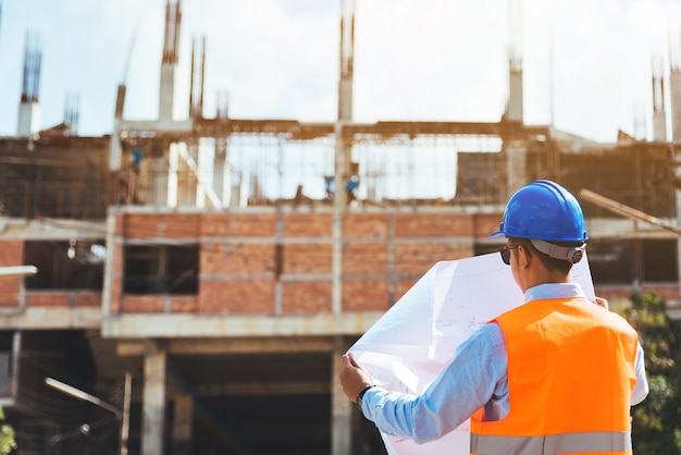 Foto vista posteriore dell'ingegnere con il casco di sicurezza blu che controlla un disegno al cantiere.