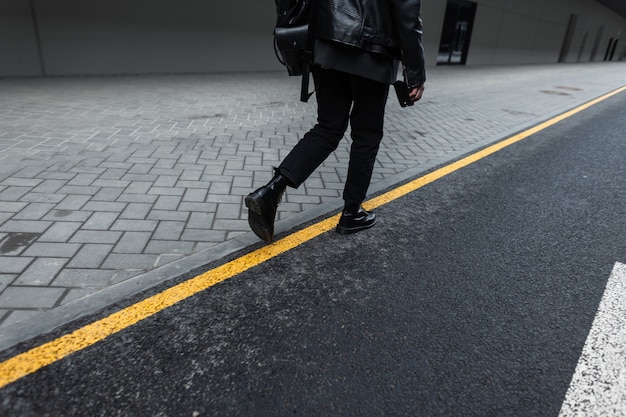 Back view on a fashionable young man in a black leather jacket in stylish jeans in leather boots with a trendy backpack in the city. Modern guy travels in fashion clothes on the street. Black style.