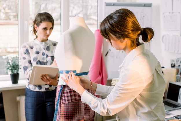 Back view of female dressmaker measuring mannequin with tape in atelier office. Talented fashion shop owner designer working with sketches for new collection of clothes. Dressmaking and sewing concept