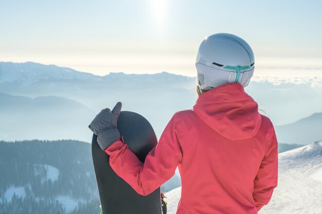 Back view of female snowboarder standing with snowboard and enjoying mountain landscape