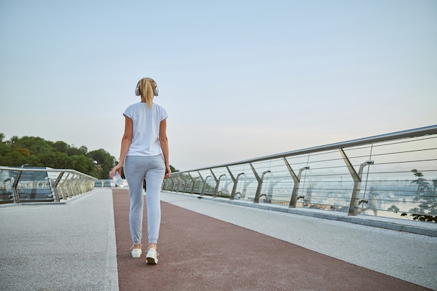 Back view of a fit blonde female with a plastic bottle walking across the bridge