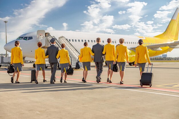 Back view of flight attendants and male pilots in work uniforms\
heading to airplane under cloudy sky at airport