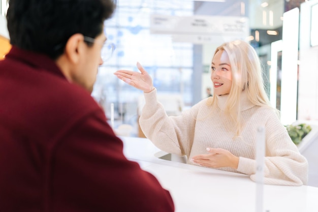 Back view from shoulder of unrecognizable man having conversation through glass partition with pretty blonde woman standing at desk in hall of shopping mall