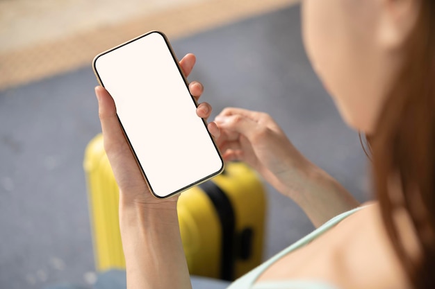 Back view of girl holding smartphone with white blank screen mockup Traveler with phone at the airport with passport and suitcase