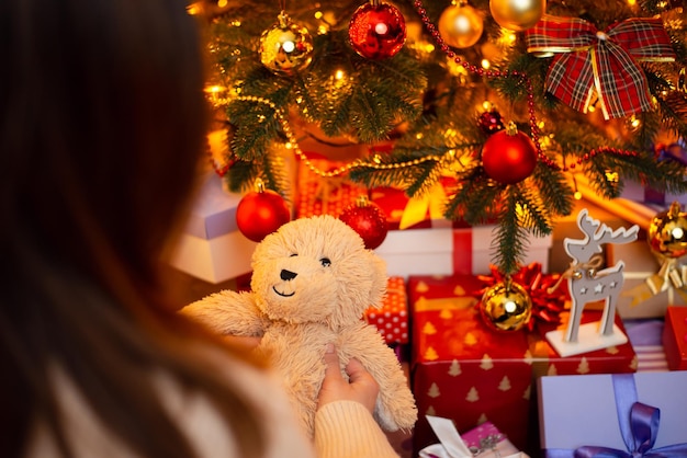 Back view of a girl holding teddy bear present on background of Christmas tree