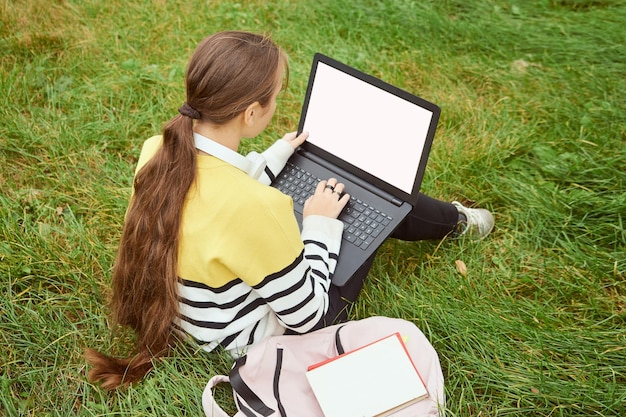 写真 後ろ姿の女子学生は、試験の準備をして芝生の上に座ってラップトップを使用しています