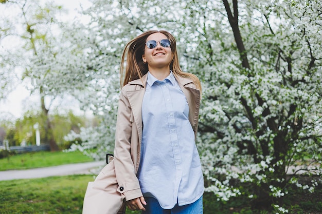 Ragazza di vista posteriore che cammina sotto il melo in fiore nel parco naturale sul pendio della collina a praga elegante giovane donna con cappello e cappotto marrone che si gode il giardino fiorito in una soleggiata giornata primaverile