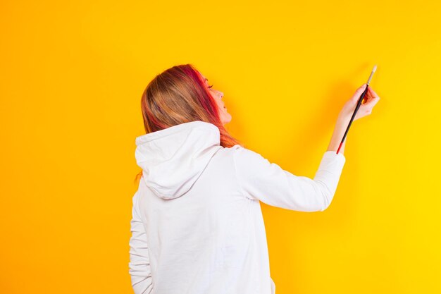 Back view of a girl who holding a brush in her hand The blonde girl in a white hoodie stands on a yellow background