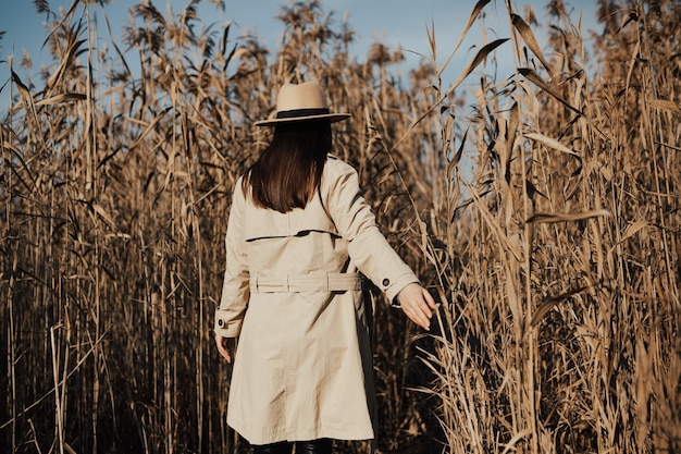 Back view of girl with beige trench coat and hat in a thicket of dry reeds with blue sky on the background.