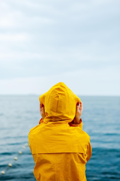 Back view of girl in yellow raincoat standing on stone pier of waterfront with ocean on background