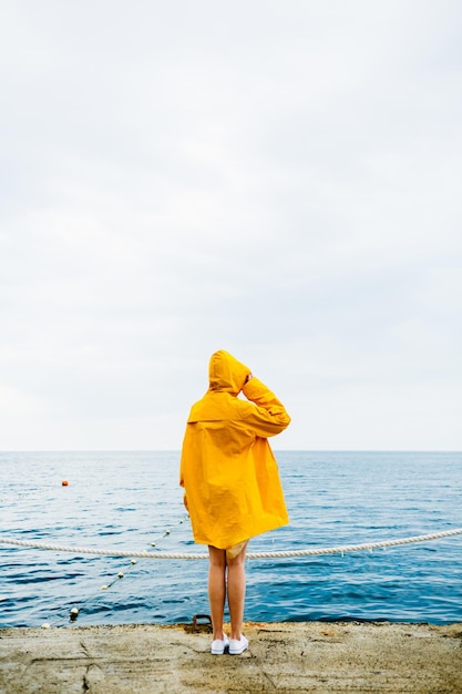 Back view of girl in yellow raincoat standing on stone pier of waterfront with ocean on background