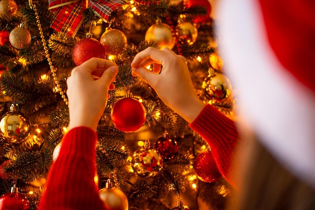 クリスマス ツリーに赤いボールをぶら下げ女の子の手の背面図 暖かい金色の光の魔法のお祭りの雰囲気