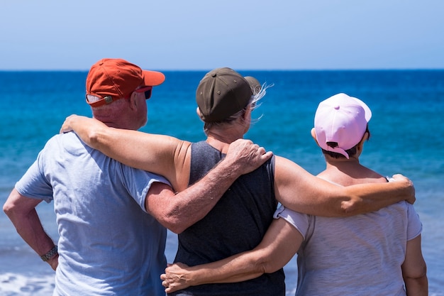 Back view of group of three friends senior people enjoying together the beach looking at the horizon over water - concept of freedom and active retired elderly  in holiday