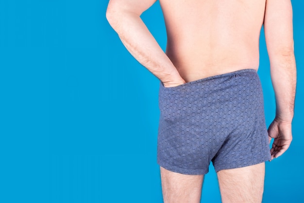 Back view of grown man in dark underwear puts his hand inside them on blue background