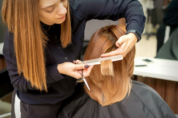 Il punto di vista posteriore del parrucchiere taglia i capelli rossi o marroni alla giovane donna nel salone di bellezza. taglio di capelli nel parrucchiere