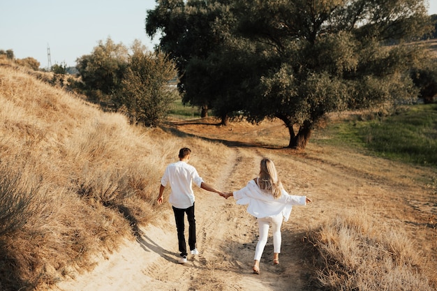 Back view of happy couple in white clothes walking and holding hands in park.