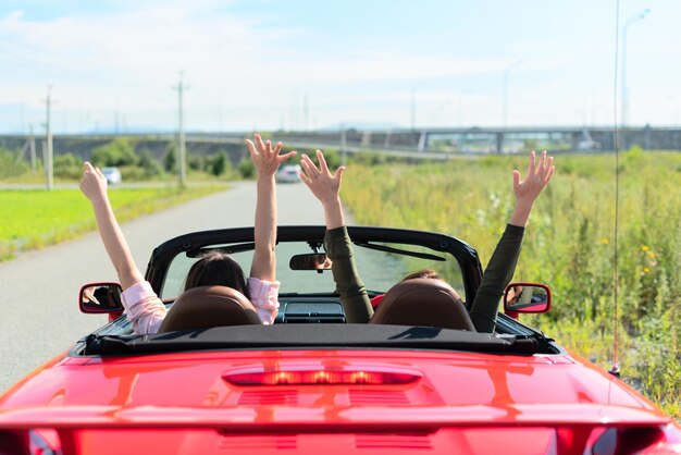 Back view of happy girls driving red cabriolet car with raised hands during vacation road trip having fun together discovering new places Road trip travel enjoying freedom concept Selective focus