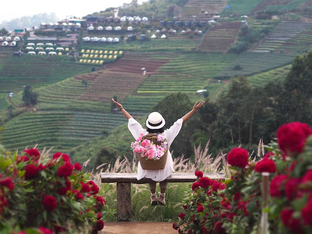 幸せな女性の観光客の背面図は、ベンチに座って、タイのチェンマイにあるモンチャムとノンホイロイヤルプロジェクトの花畑で腕を上げます。