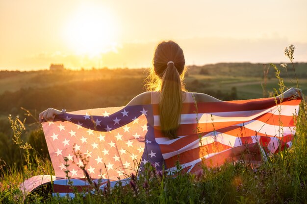 Back view of happy woman with USA national flag sitting outdoors at sunset. Positive girl celebrating United States independence day. International day of democracy concept.