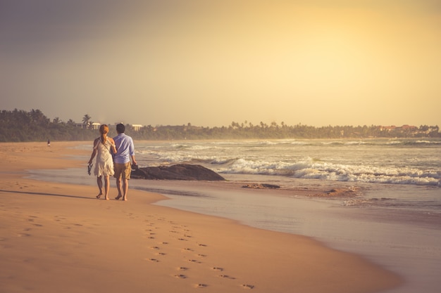 人けのない熱帯のビーチの上を歩いて幸せな若いカップルの背面図です。調色写真