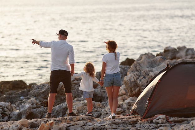 Foto punto di vista posteriore di giovani genitori felici, tenendosi per mano figlia, stante sulla spiaggia della roccia.