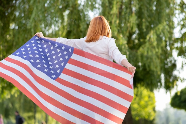 back view of happy young red haired woman posing with usa national flag standing outdoors