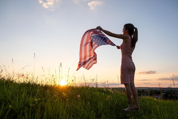 Back view of happy young woman posing with USA national flag outdoors at sunset Positive female celebrating United States independence day International day of democracy concept